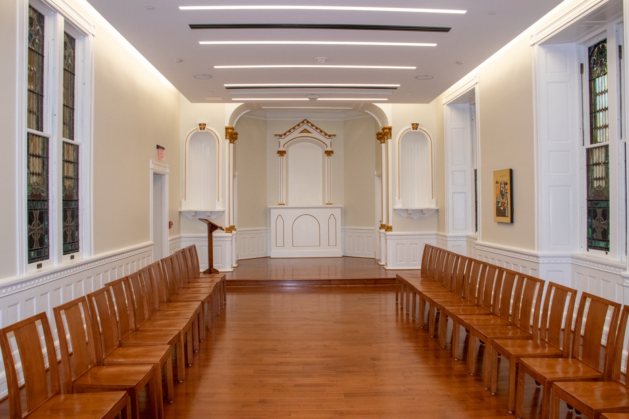 Picture of the ecumenical chapel. Orange colored wood flooring with two rows of chairs on either sided of the image. There is an altar in the back of the image with stained glass windows on either sided, and a painting of St. Ignatius on the left wall.