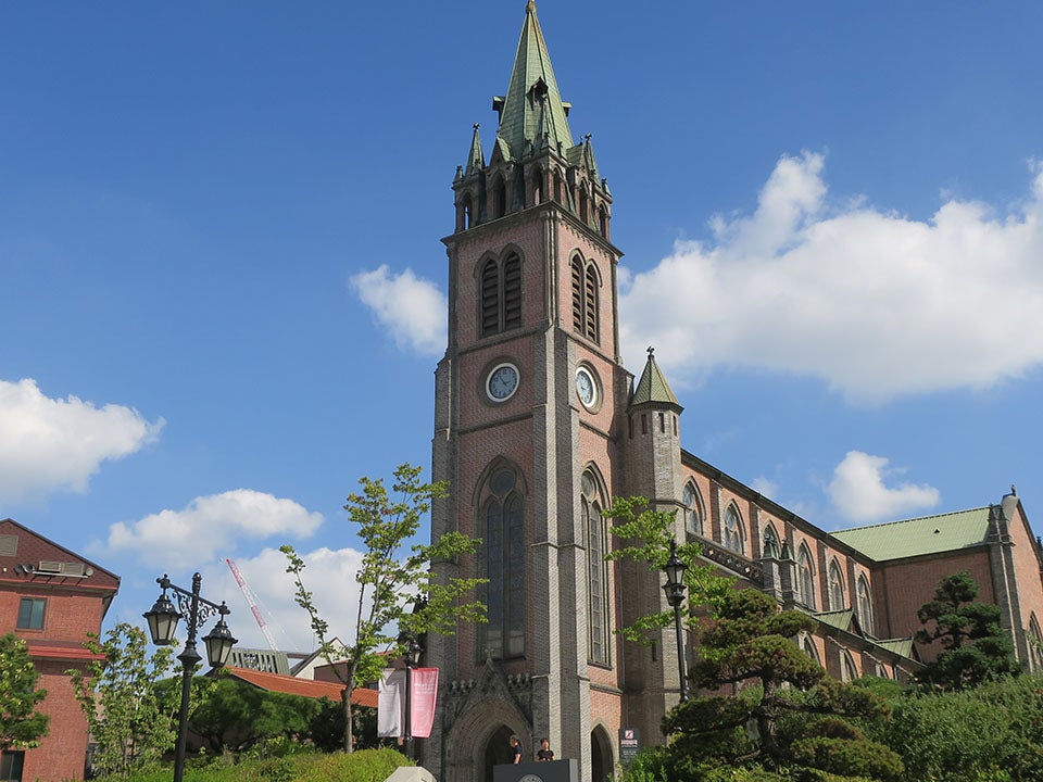 A Cathedral clock tower in the foreground surrounded by trees and shrubs. The Cathedral Church of Our Lady of the Immaculate Conception in Seoul, Korea