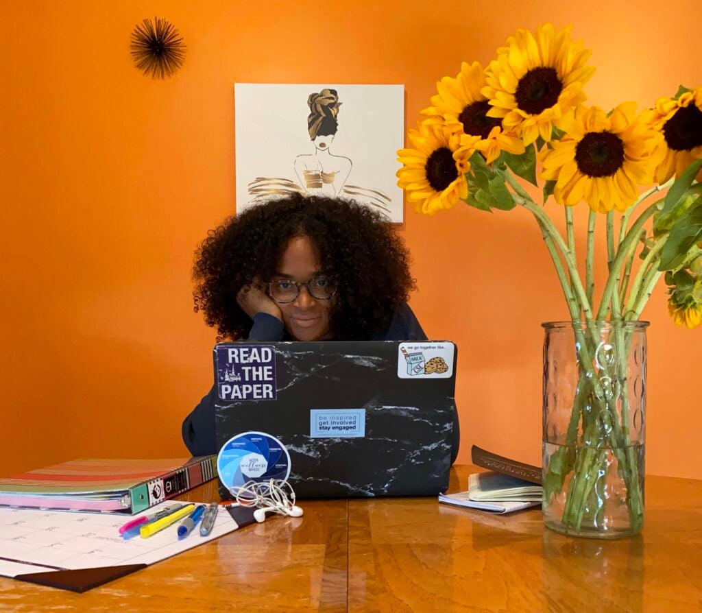 Jaime is sitting at a table with her head peeking out from behind a laptop, with curly dark hair and a hand on her cheek. She’s in a bright orange room. Her laptop has several stickers on it including the Georgetown Wellness Wheel. She’s wearing glasses and a gray sweater. On the table are a vase of long stem sunflowers, a binder, a calendar and colorful pens. 