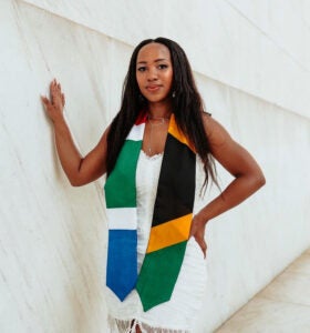 A Black woman is standing next to a white marble wall wearing a white dress with a stole in the colors of South Africa.