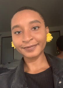 portrait style photo of a young Black woman. She is wearing a black t-shirt and jacket and wearing yellow earings.
