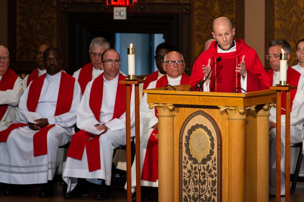priests in white robes with red stoles, seated behind a presider, also a priest in red vestments standing at a podium speaking