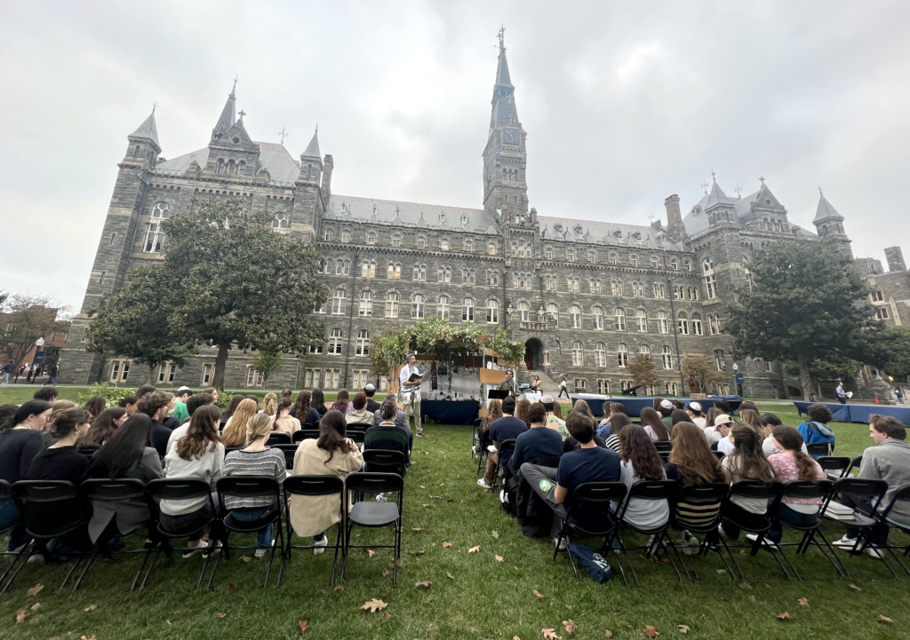 People seated in chairs arranged in rows facing the a rabbi delivering a sermon with Healy Hall, a Victorian Gothic-style building.