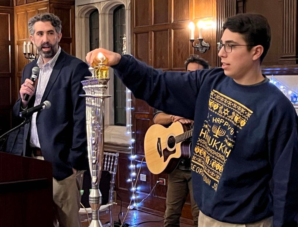 A bespectacled male student wearing a blue sweatshirt lights a candle on a menorah while a man with a beard, a rabbi recites the blessings for lighting the Hanukkah candles