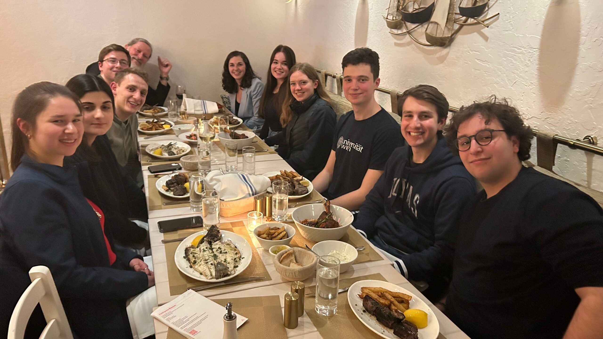 Nine students and trip chaperones, Fr Pratt and Becky Hay sitting at a dining table with plates of food in front of each person. 