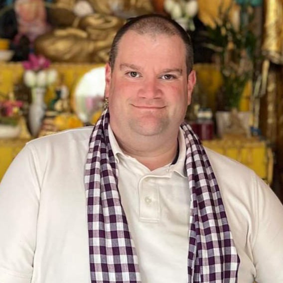 A white man with shaved head wearing a cream colored shirt and checked scarf. 