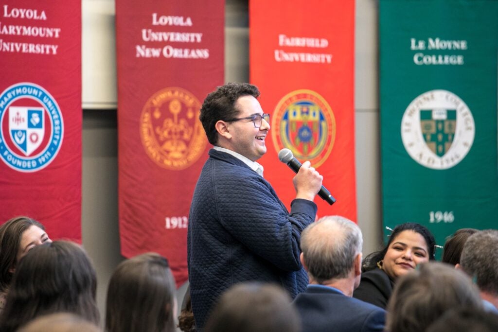 A man wearing glasses and a blue jacket speaking into a microphone among a group of people. Colored banners in the background. 