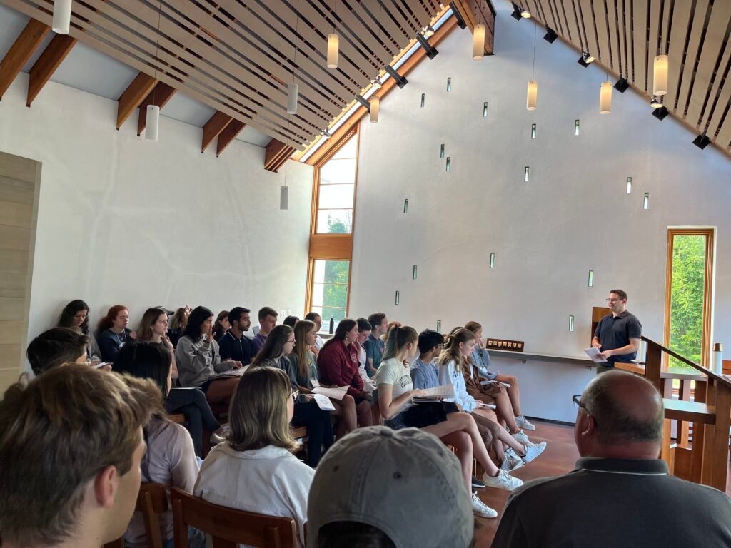 A group of students seated in rows inside a chapel. Attention is focused on a speaker, a man wearing a blue shirt, and glasses. 