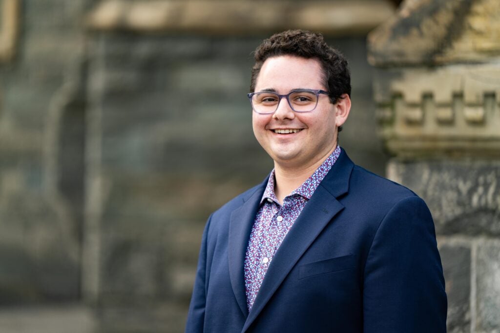 A professional photo of a man, wearing a blue blazer and glasses. He is standing in front of a stone wall.