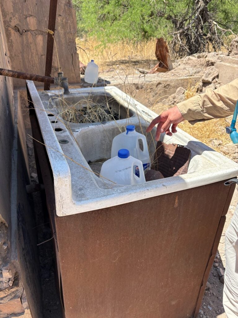 a hand is pointing out jugs of water left in an old sink.