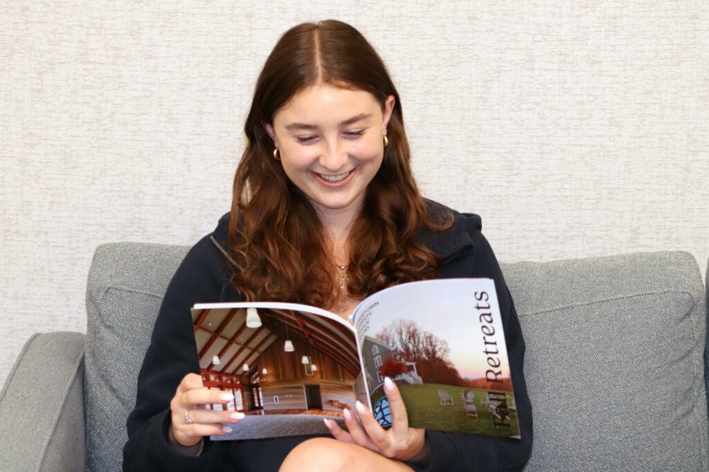 A young woman with long red hair is sitting on a grey couch. she is holding a magazine and smiling.