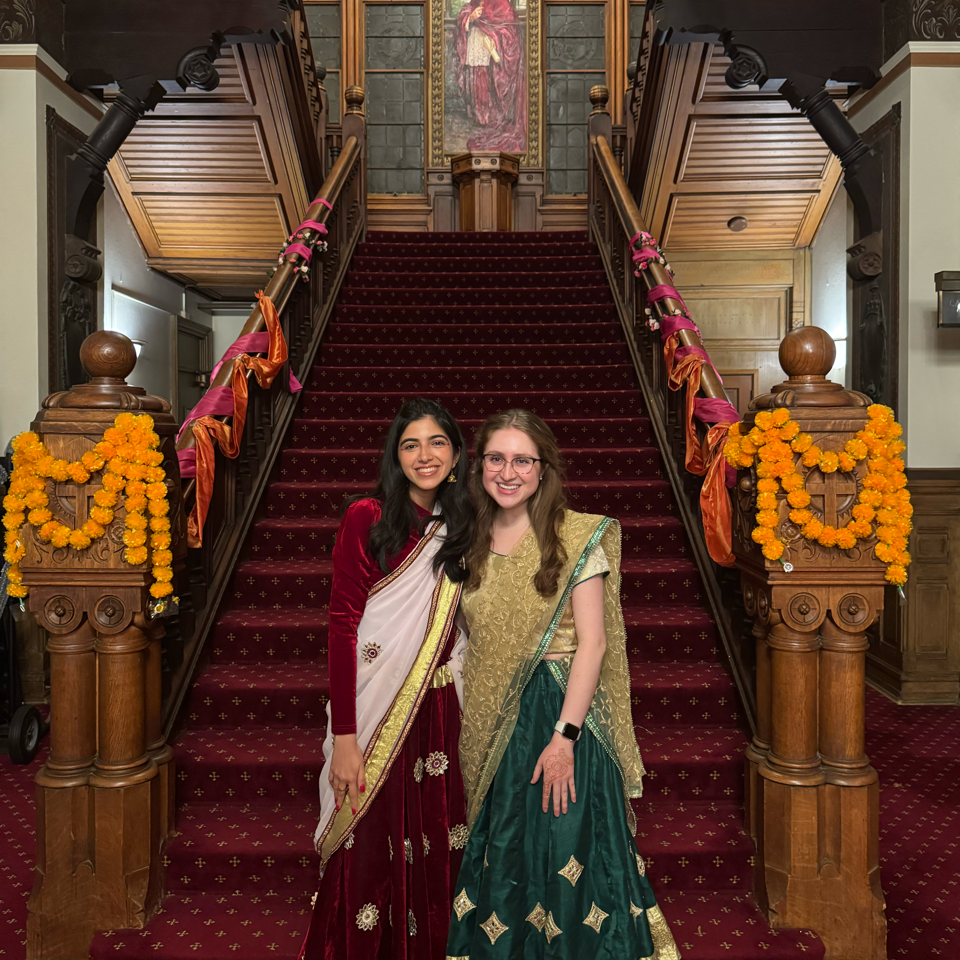 Saara Bidiwala (C’26) and Claire Auslander (N’26) dressed in sarees, standing in front of a staircase.