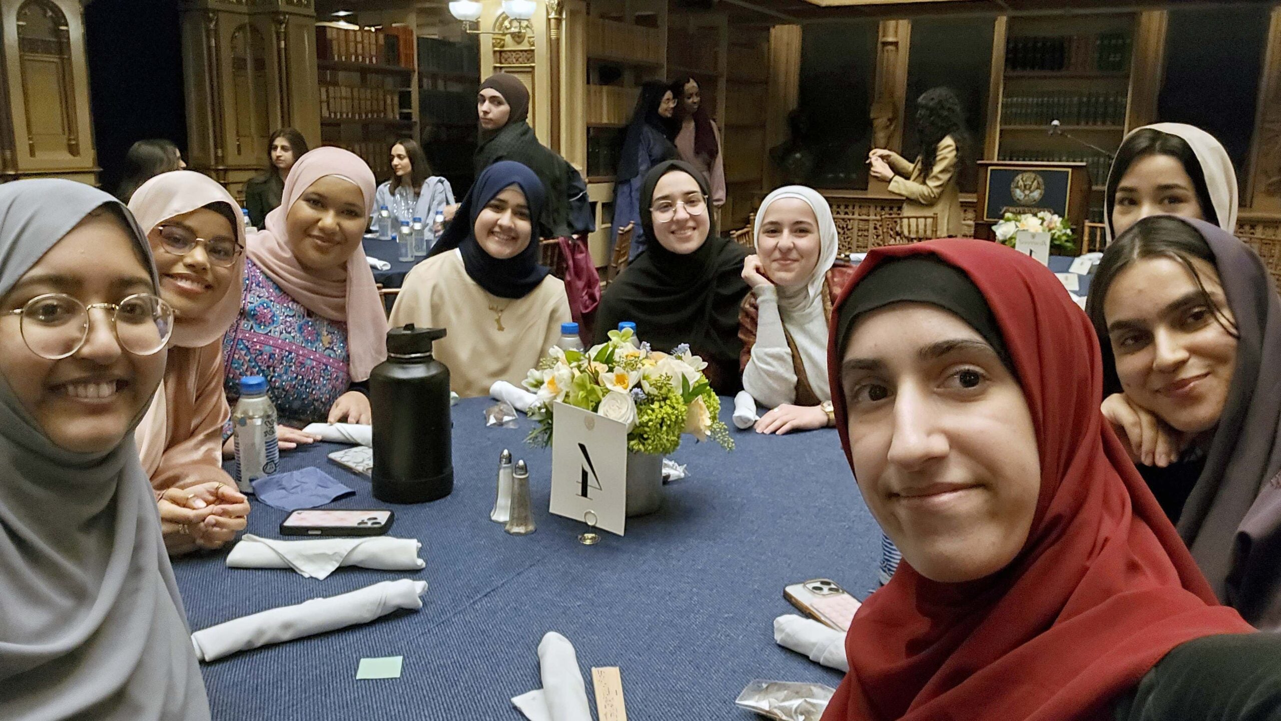 a group of young women wear head scarves sits around a table.