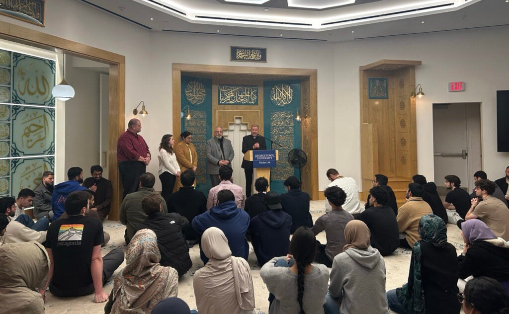 A group of muslim students sit on a floor facing five speakers at the front of the room.