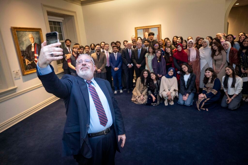 A bearded man standing with his back to a group of people is attempting to take a selfie.
