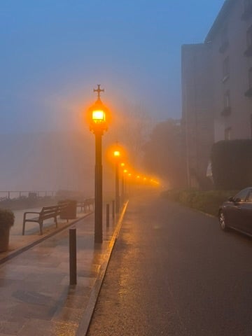 A street full of fog and warm, glowing orange lampposts and benches on the left side. 