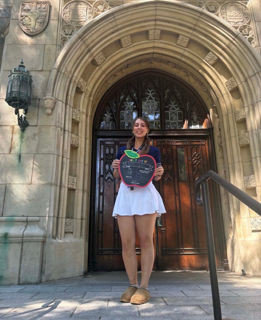 A young woman is standing in front of a doorway holding an apple-shaped sign that reads "first day of college." 