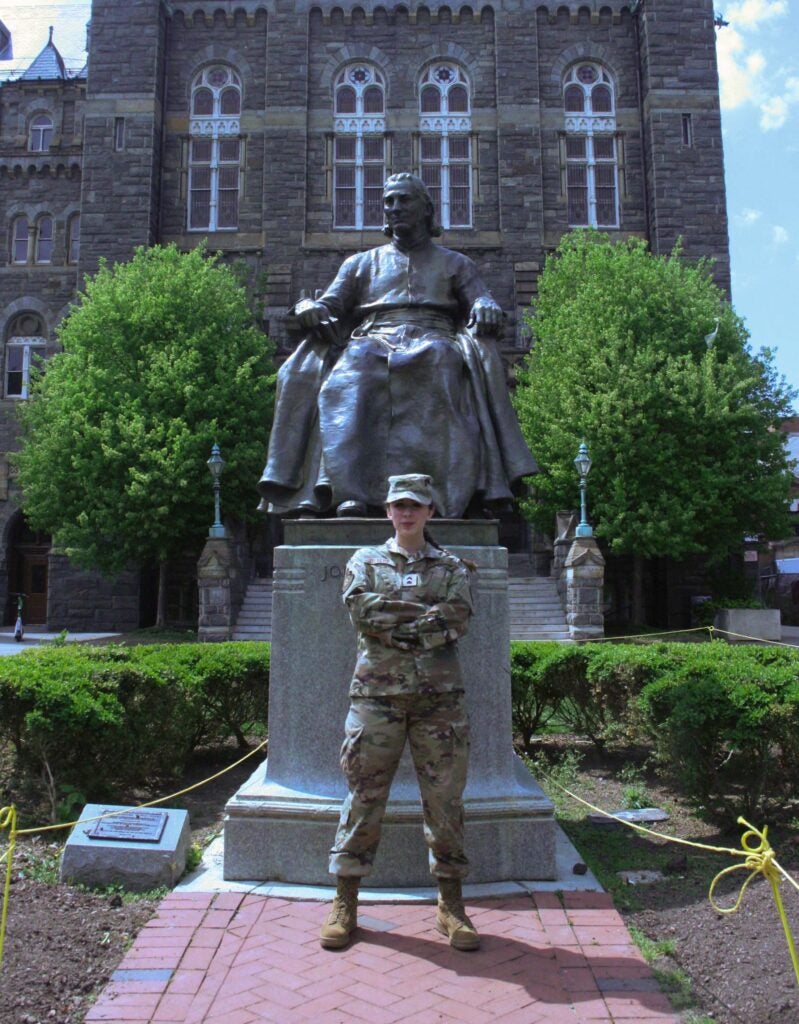 A woman in army fatigues poses in front of a statue of a priest. 