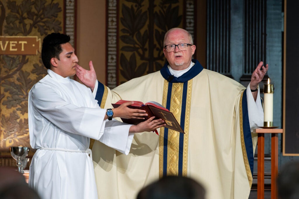 A young man with dark hair dressed in a white robe is holding a Bible for a priest during a mass service. 