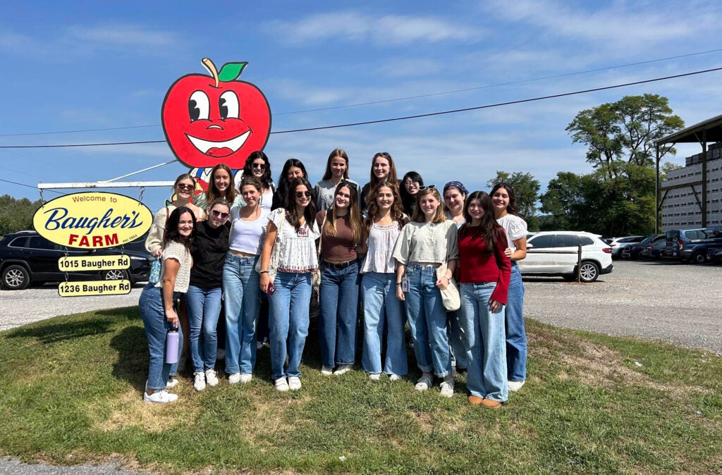 A group of young women stand together outside. Behind them is a tall sign in the shape of a red apple. 