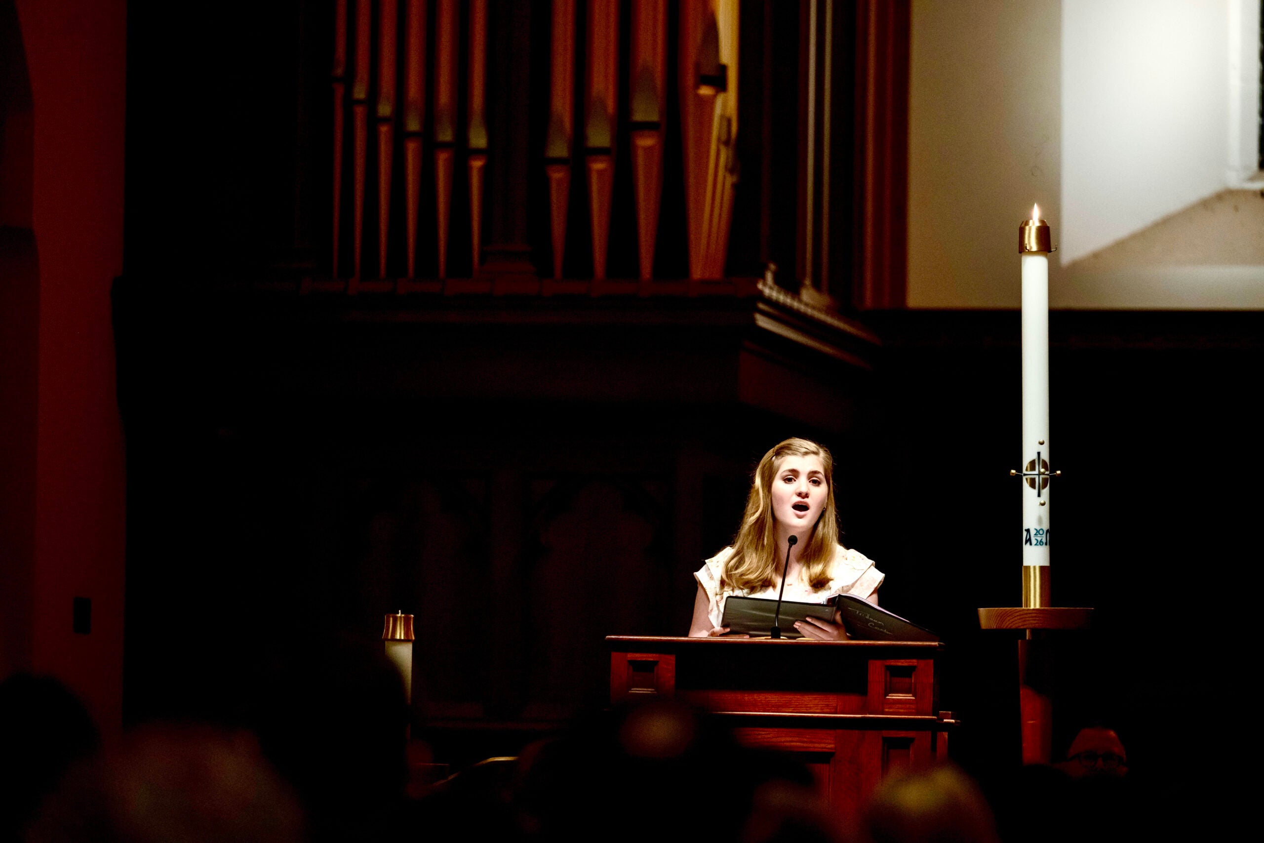 A young woman with blond hair stands at a lecturn, inside a chapel, singing.