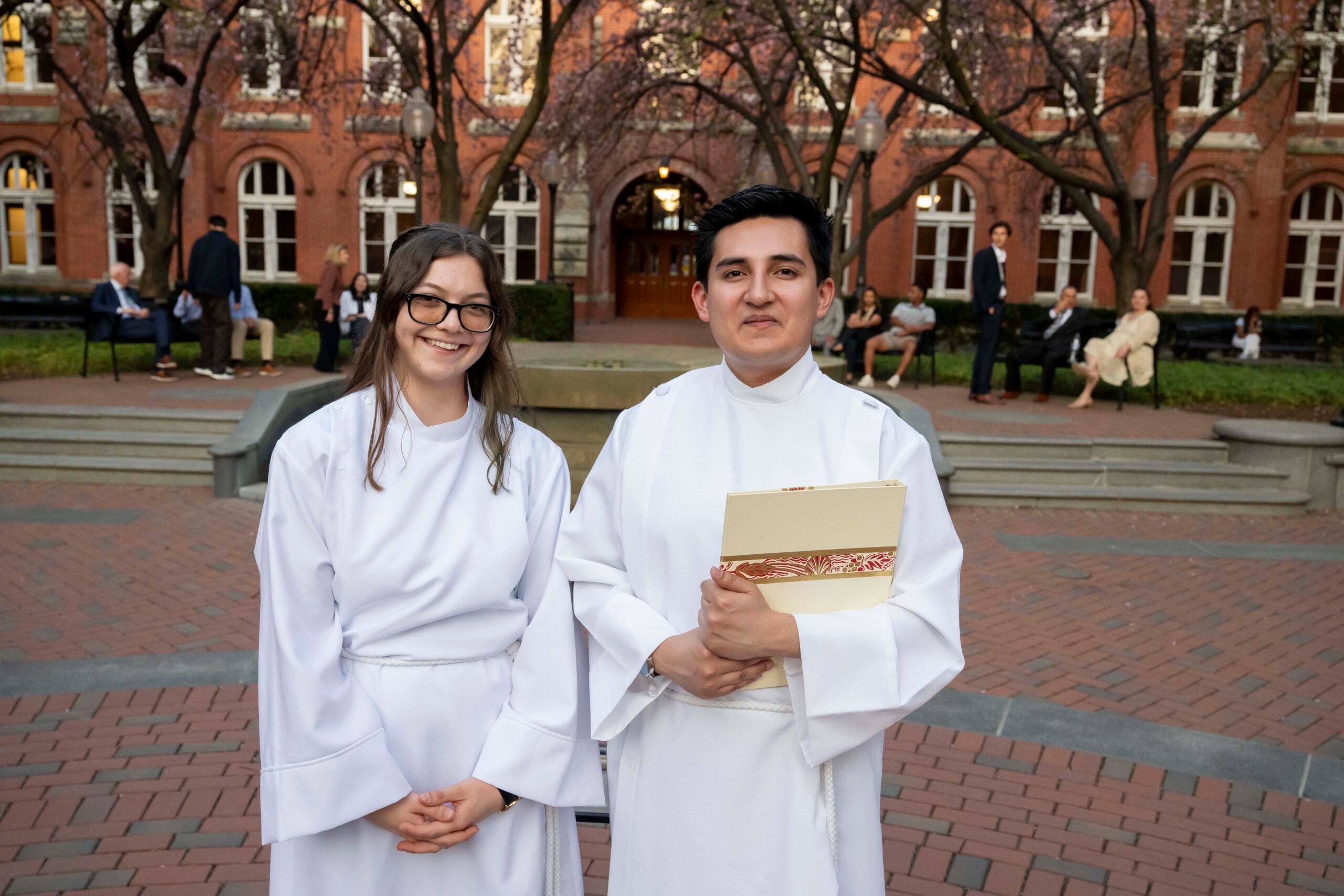 a young woman and young man, both wearing white robes are standing in a cobblestone courtyard.