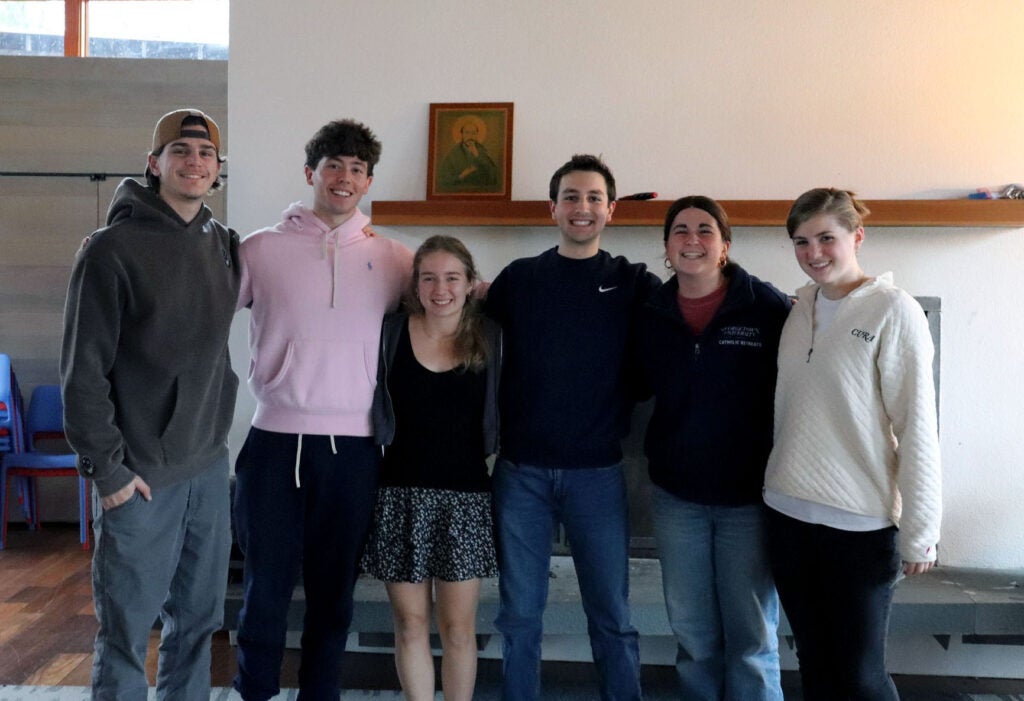 Six young people, three male and three female stand together with arms around each other.