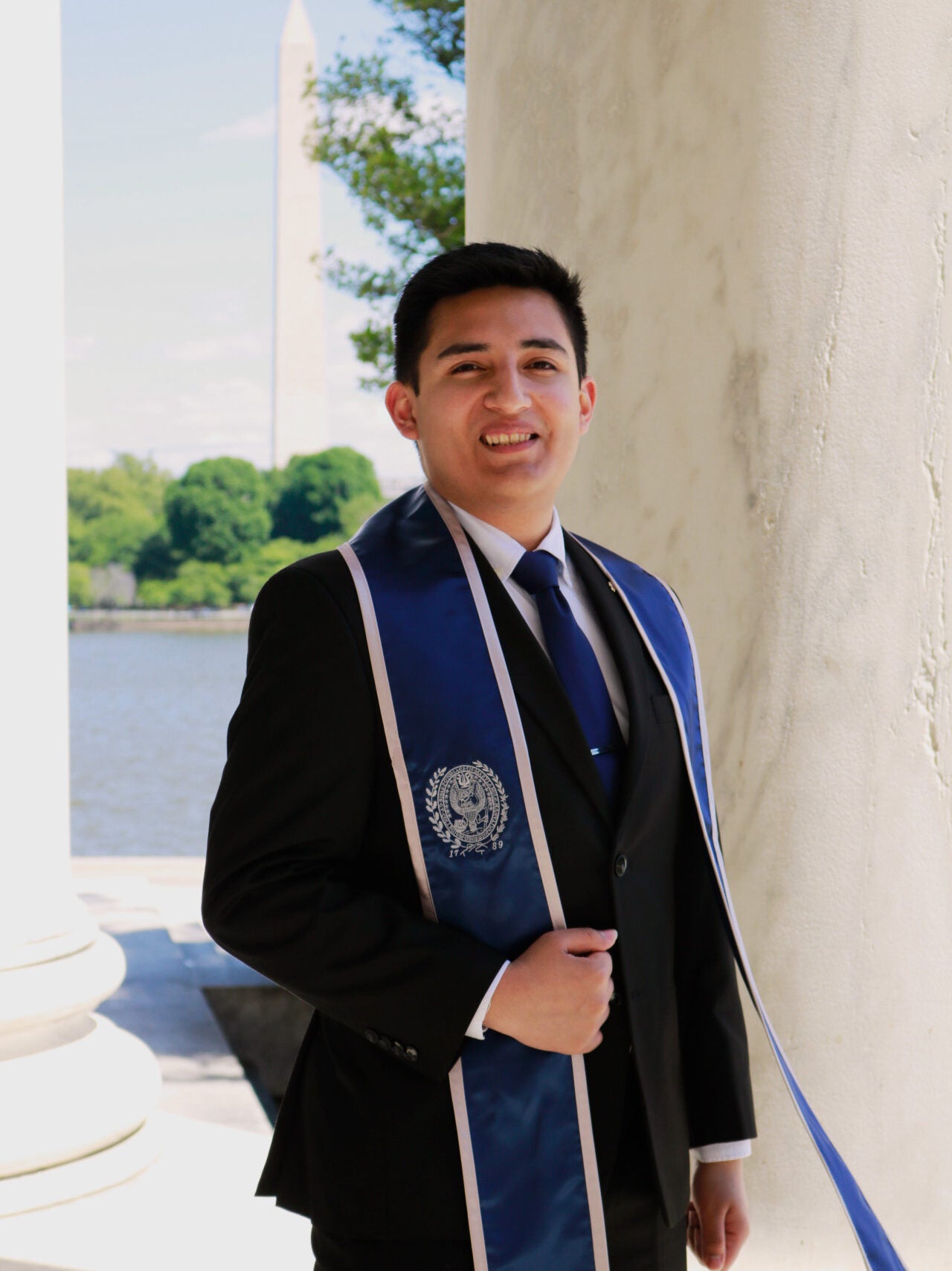 A young man with dark hair, wearing a dark suit with a white shirt and blue tie and a blue graduation stole is standing beside a marble column. 