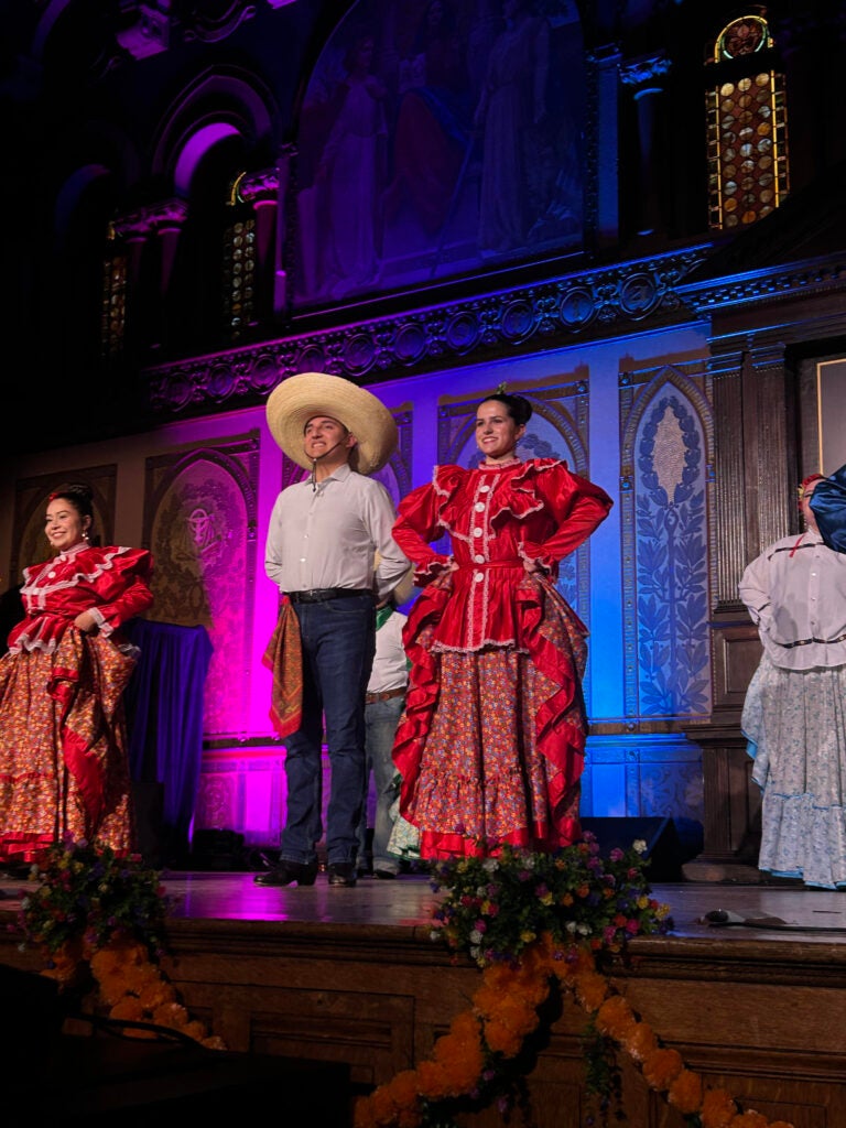 Dancers in the traditional dress of Mexico's folk ballet are standing on a stage lit with purple light.