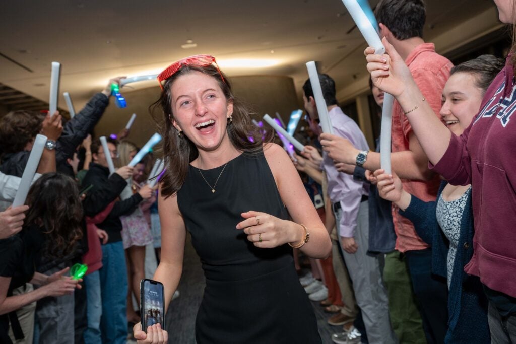 a young woman wearing a black dress with red sunglasses tucked in her hair is smiling and surrounded by other people waving celebration sticks.