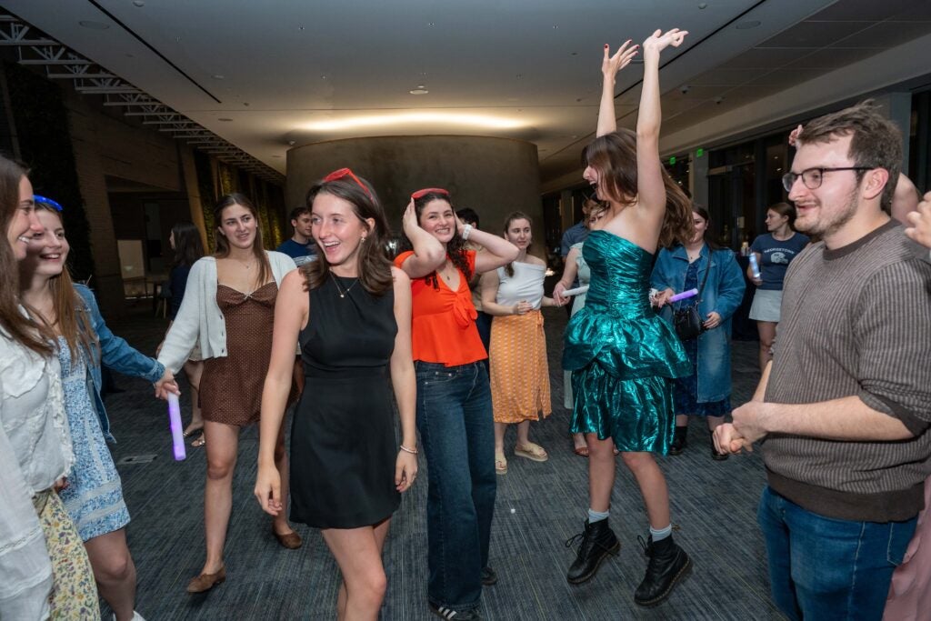 a group of young people are dancing, one of the women in a shiny green dress is jumping with her hands in the air.