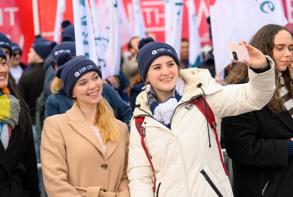 Two young women, both wearing blue beanie hats, and light colored winter jackets. One woman is holding up a smartphone to take their picture. 