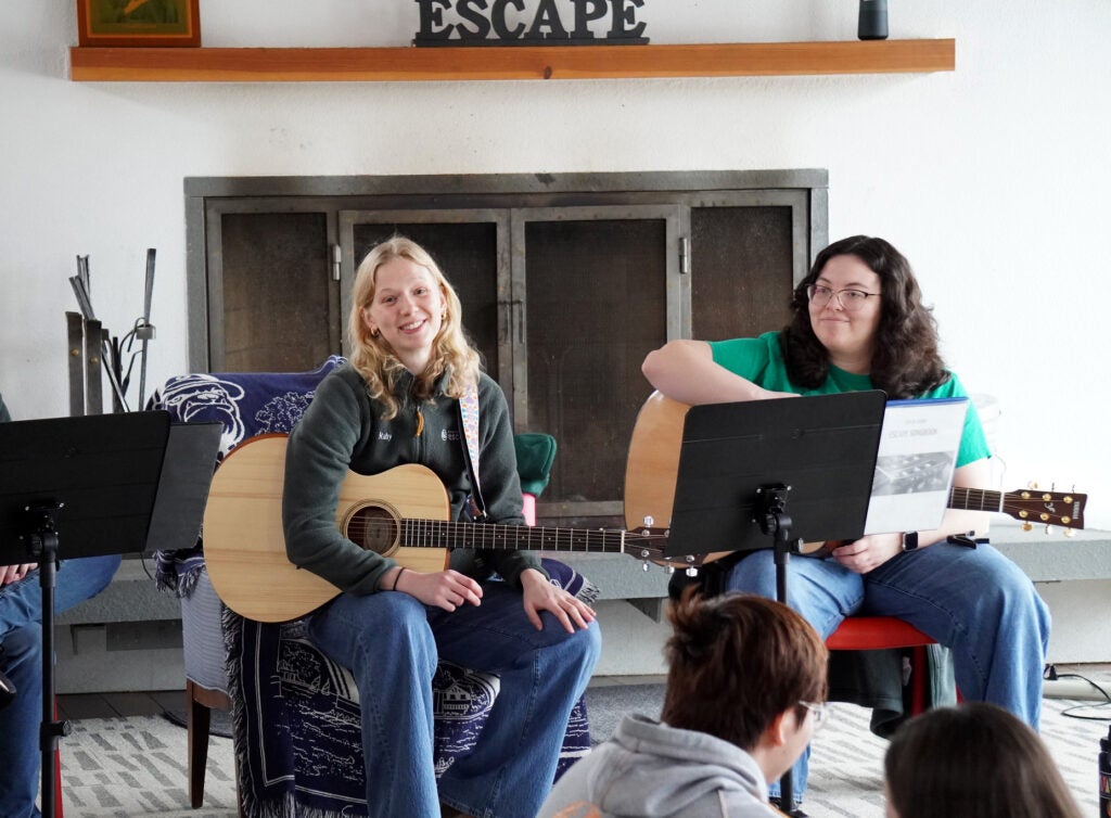 Two young women, sitting on chairs each has a guitar on her lap. They are leading a sing along for students. 