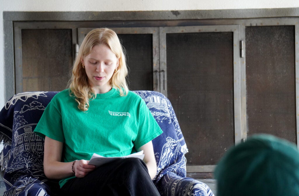 A young woman, blond hair, wearing a green T-shirt, dark trousers, is giving a talk.