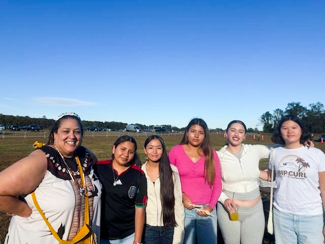 Six indigenous women, one in traditional garments outdoors at a festival.