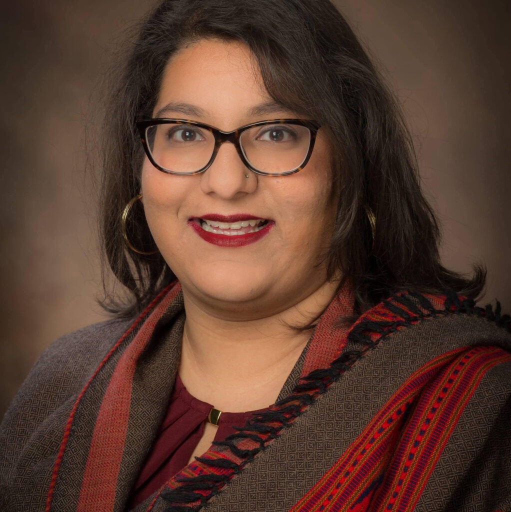 A woman with shoulder-length brown hair, wearing eye glasses and a brown and red scarf.