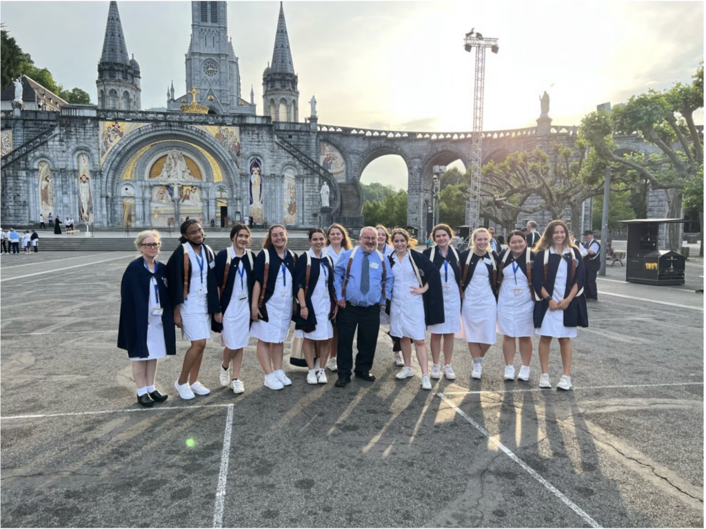 Nursing students dressed in white uniforms with blue capes stand in front of an arched gateway in Lourdes.