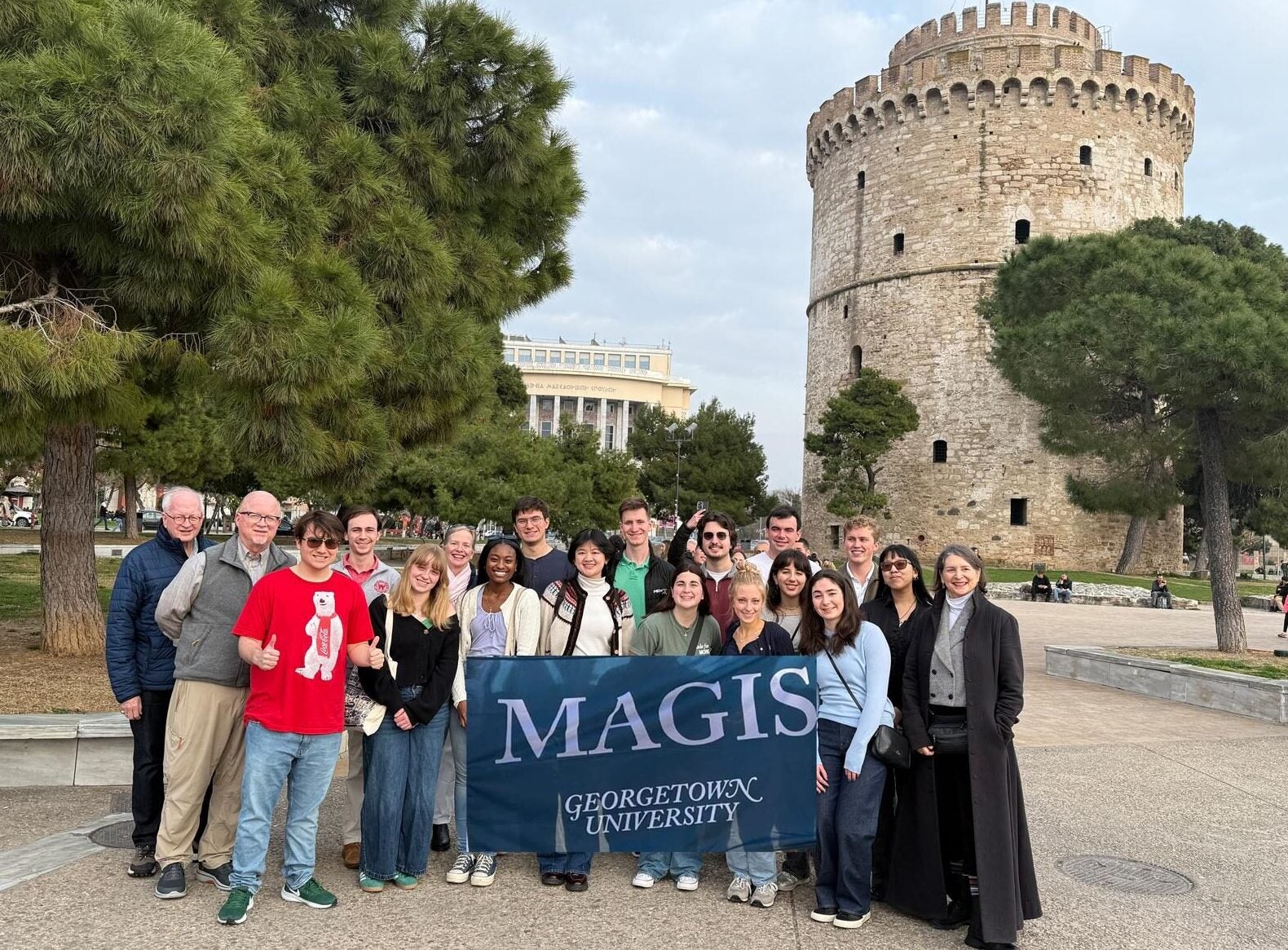 A group of people holding a "Magis" flag are standing in front of large pine trees and an ancient tower.